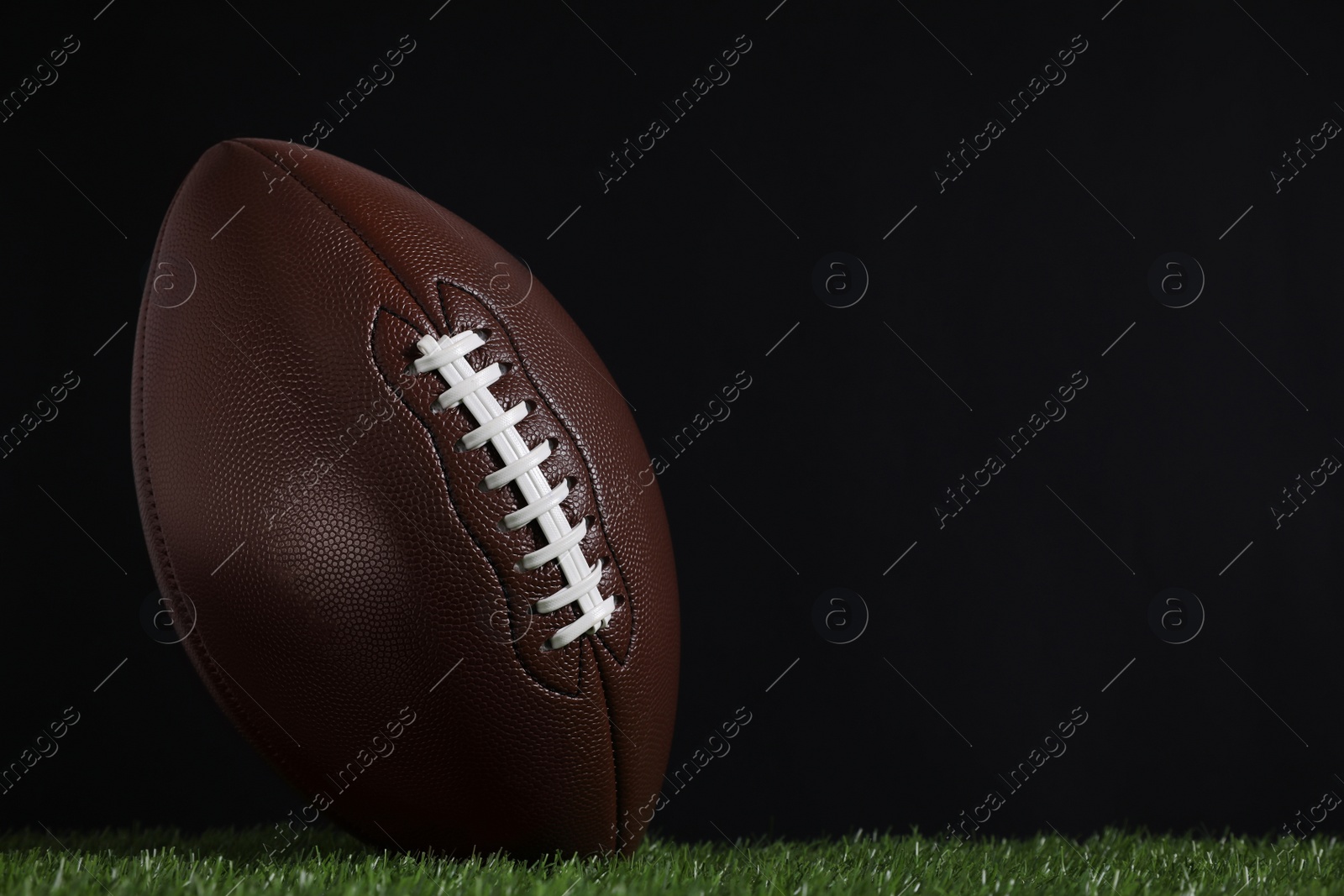 Photo of American football ball on green grass against black background. Space for text