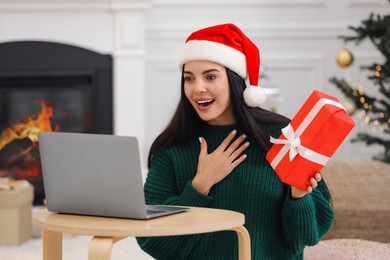 Photo of Celebrating Christmas online with exchanged by mail presents. Surprised woman in Santa hat thanking for gift during video call at home
