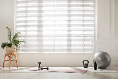 Photo of Exercise mat, dumbbells, kettlebell, fitness ball and bottle near window in spacious room