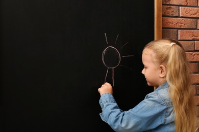 Photo of Cute little left-handed girl drawing on chalkboard near brick wall