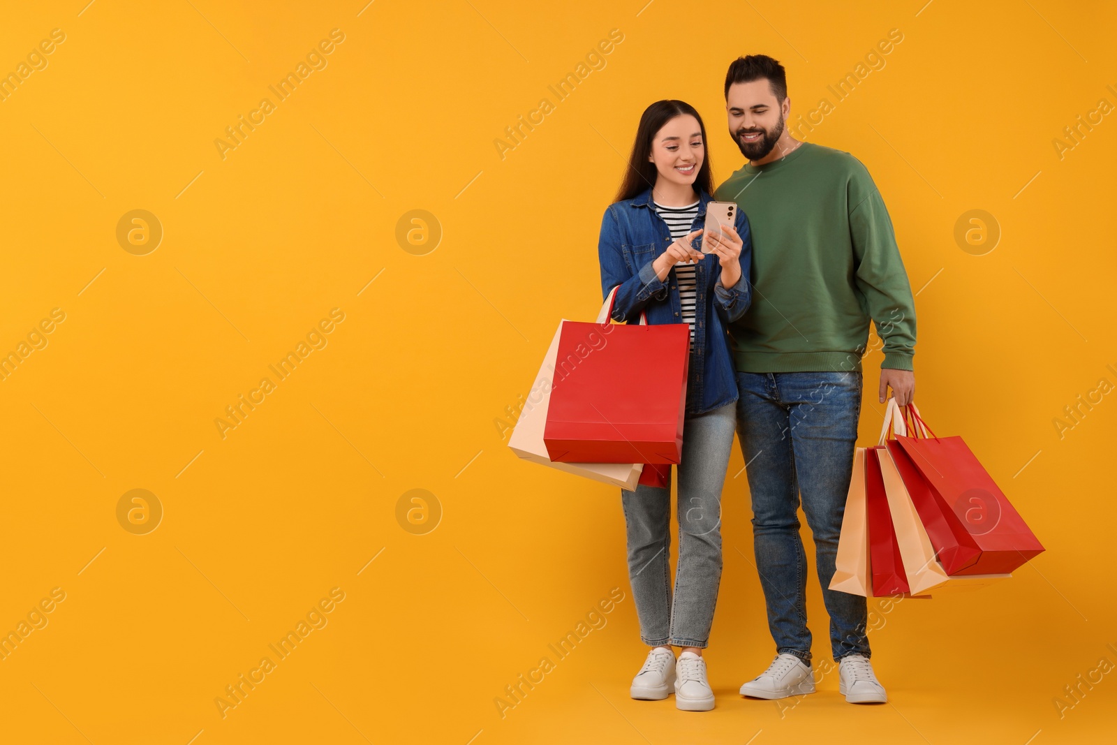 Photo of Happy couple with shopping bags and smartphone on orange background. Space for text