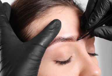 Photo of Beautician tweezing woman's eyebrow on light background, closeup