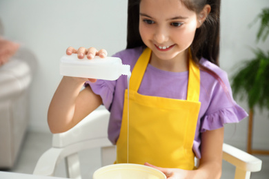 Cute little girl pouring glue into bowl at table in room. DIY slime toy