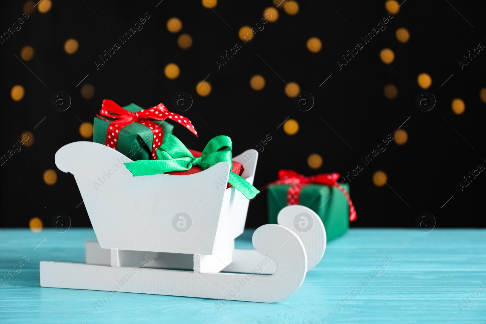 Photo of Sleigh with gift boxes on turquoise wooden table against blurred lights