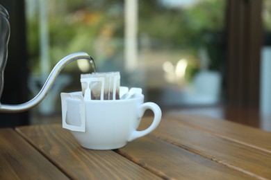 Photo of Pouring hot water into cup with drip coffee bag from kettle on wooden table
