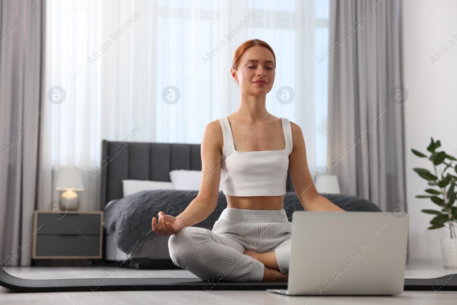 Photo of Beautiful young woman practicing Padmasana with laptop on yoga mat at home, low angle view. Lotus pose