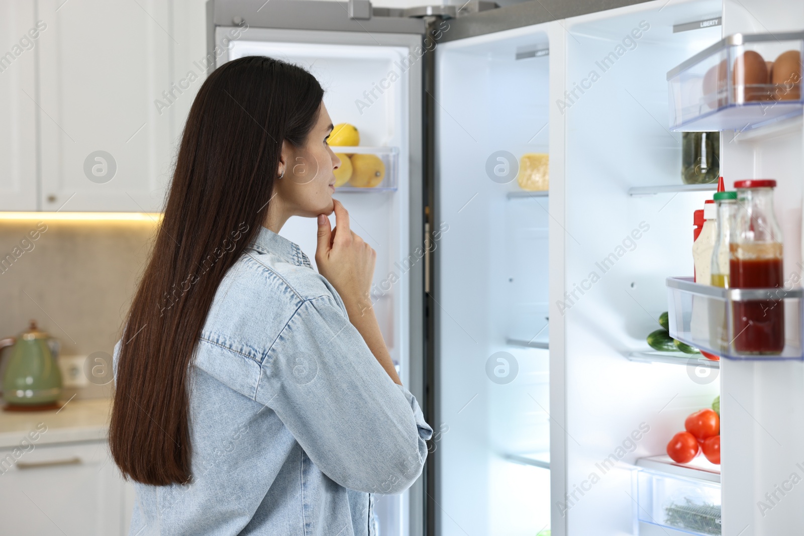 Photo of Thoughtful young woman near modern refrigerator in kitchen