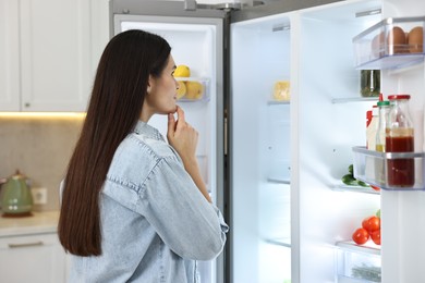 Photo of Thoughtful young woman near modern refrigerator in kitchen