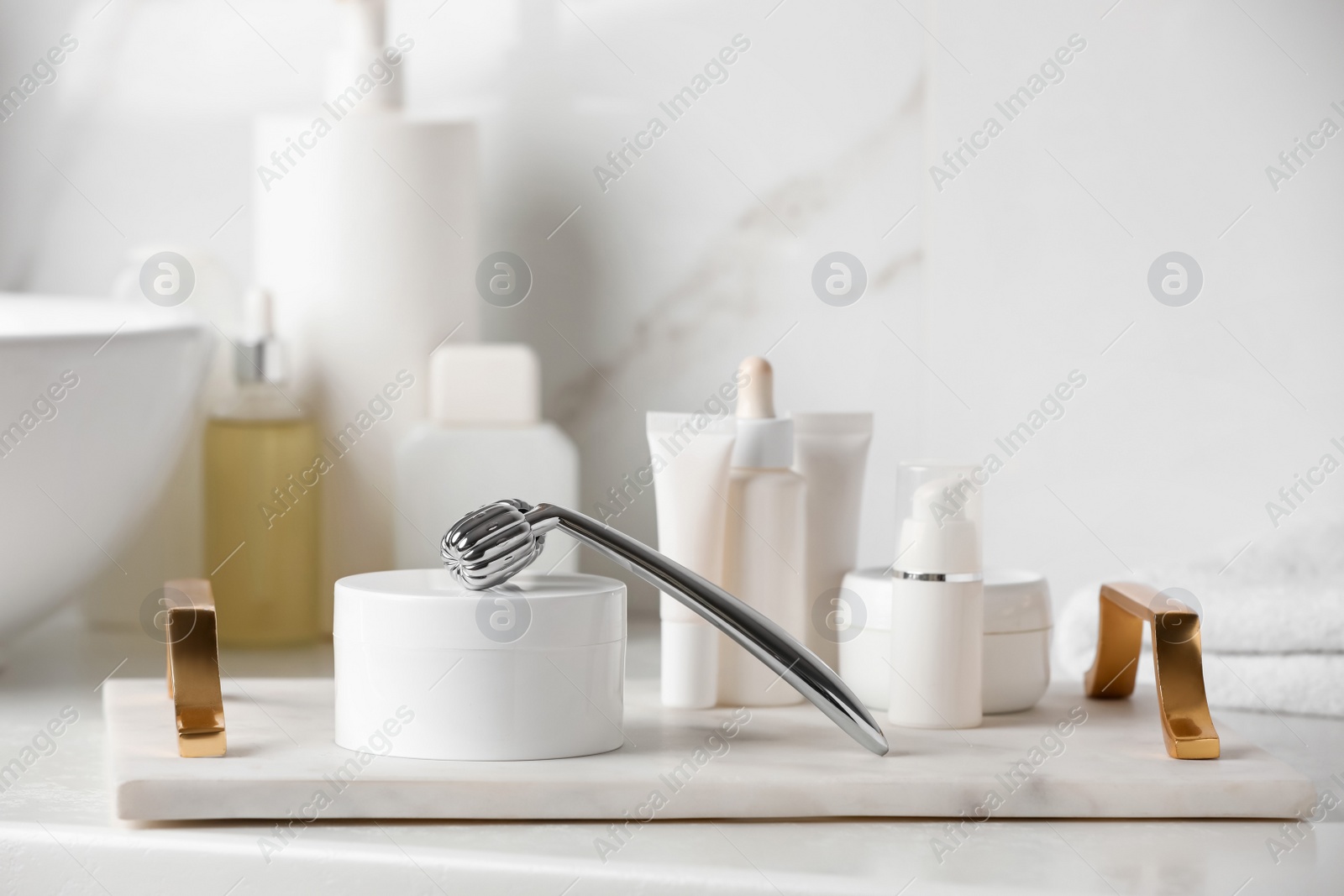 Photo of Tray with metal face roller and cosmetic products on counter in bathroom