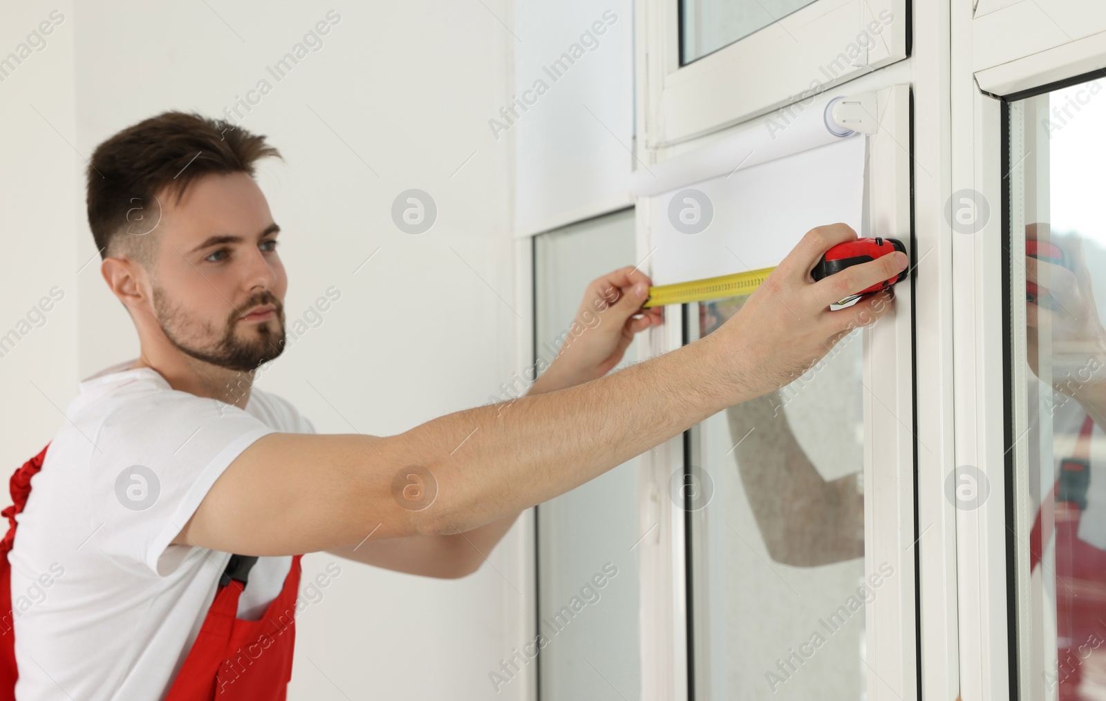 Photo of Worker in uniform using tape measure while installing roller window blind indoors, focus on hands