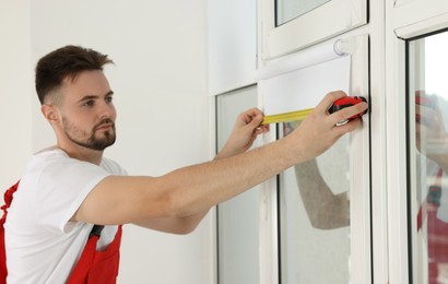 Photo of Worker in uniform using tape measure while installing roller window blind indoors, focus on hands