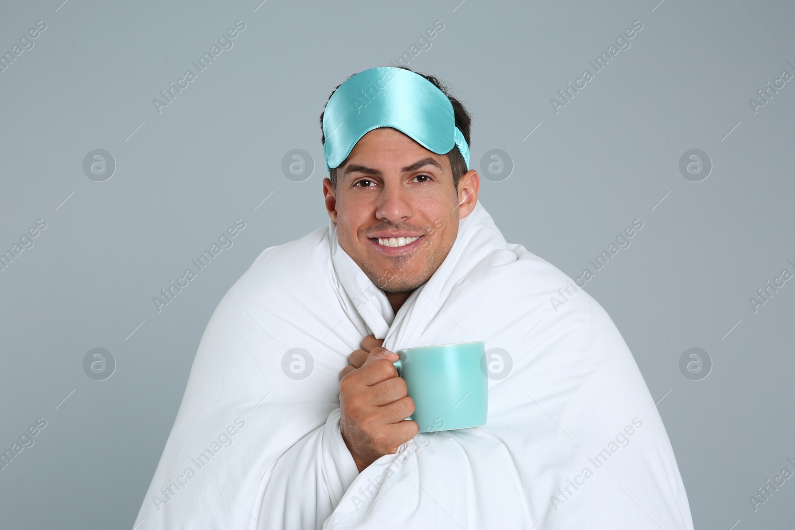 Photo of Man in sleeping mask wrapped with blanket holding cup on grey background