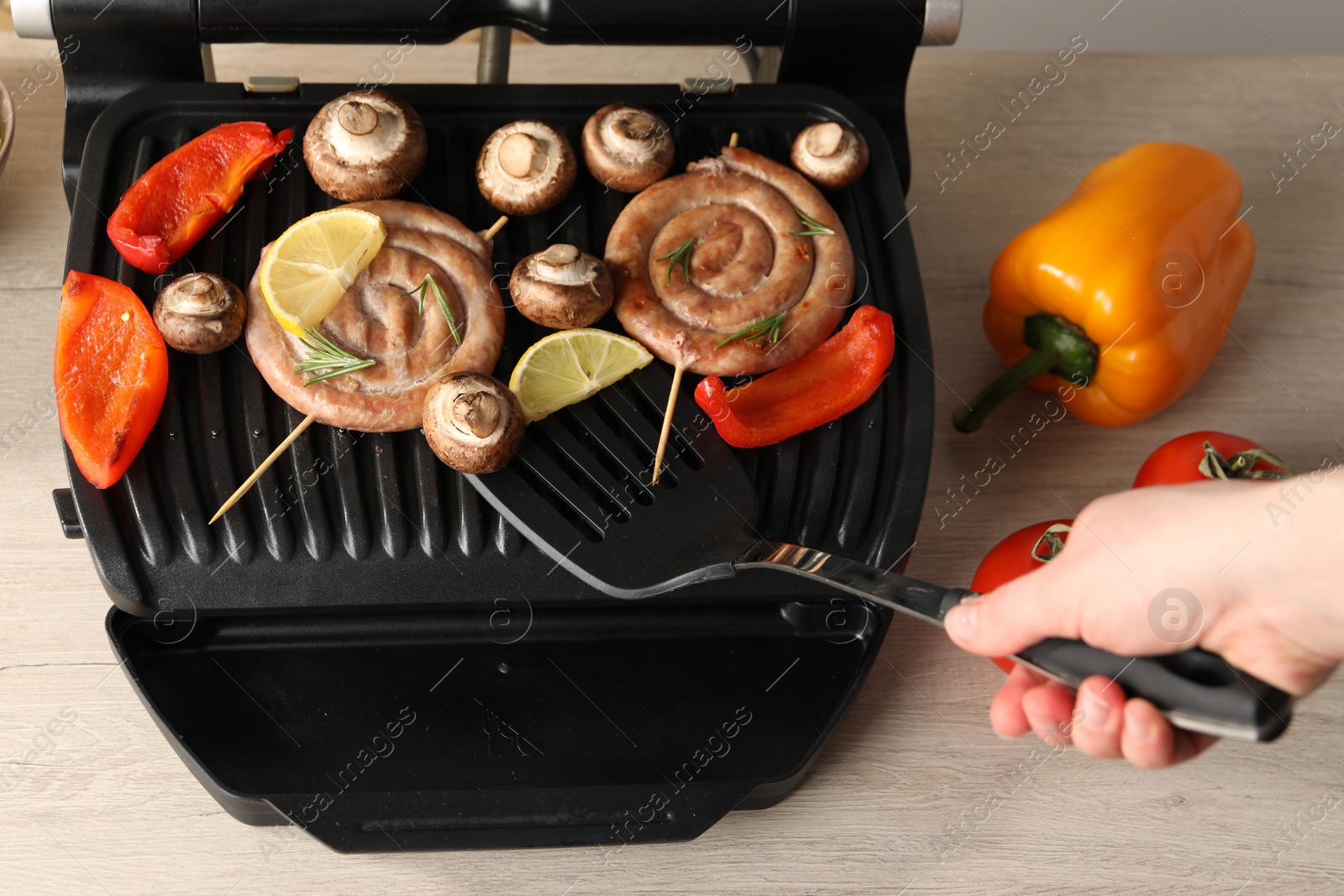 Photo of Woman cooking homemade sausages with bell peppers and mushrooms on electric grill at wooden table, closeup