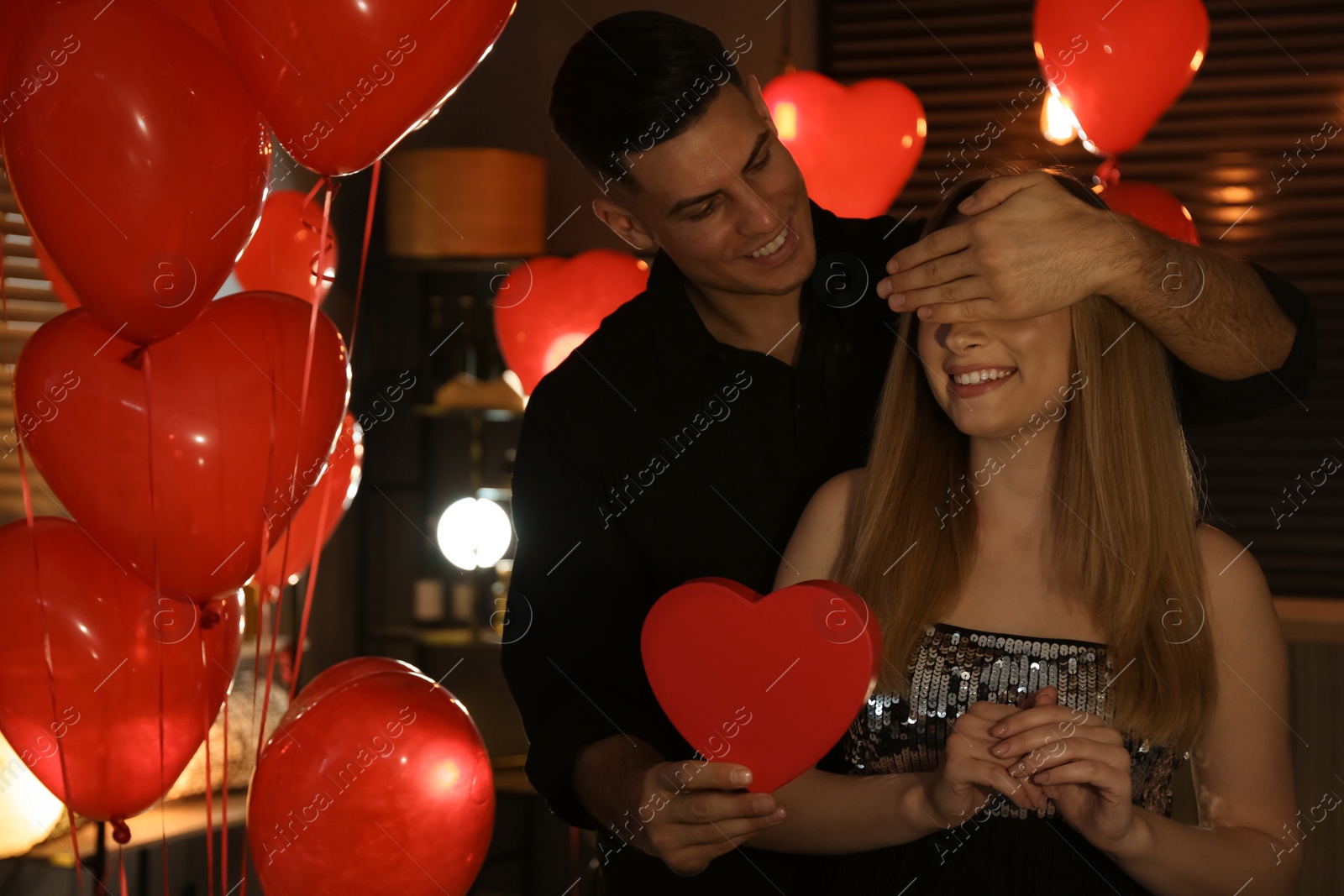 Photo of Man presenting gift to his girlfriend in room decorated with heart shaped balloons. Valentine's day celebration