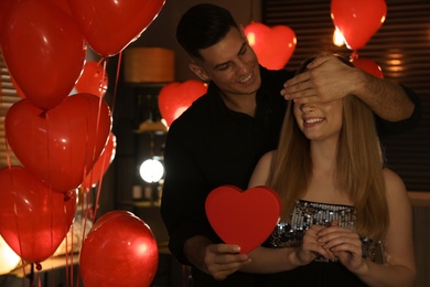 Man presenting gift to his girlfriend in room decorated with heart shaped balloons. Valentine's day celebration