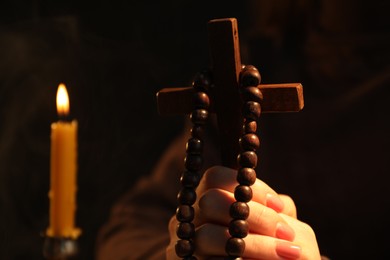 Photo of Woman with cross, beads and burning candle praying on blurred background, closeup