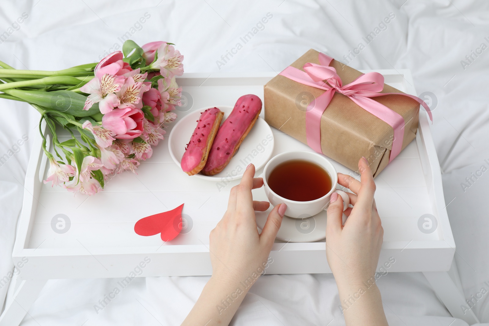 Photo of Tasty breakfast served in bed. Woman with tea, eclairs, gift box and flowers at home, closeup