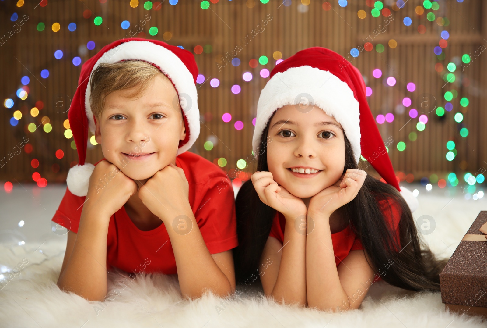 Photo of Cute little children in Santa hats lying on floor against blurred lights. Christmas celebration