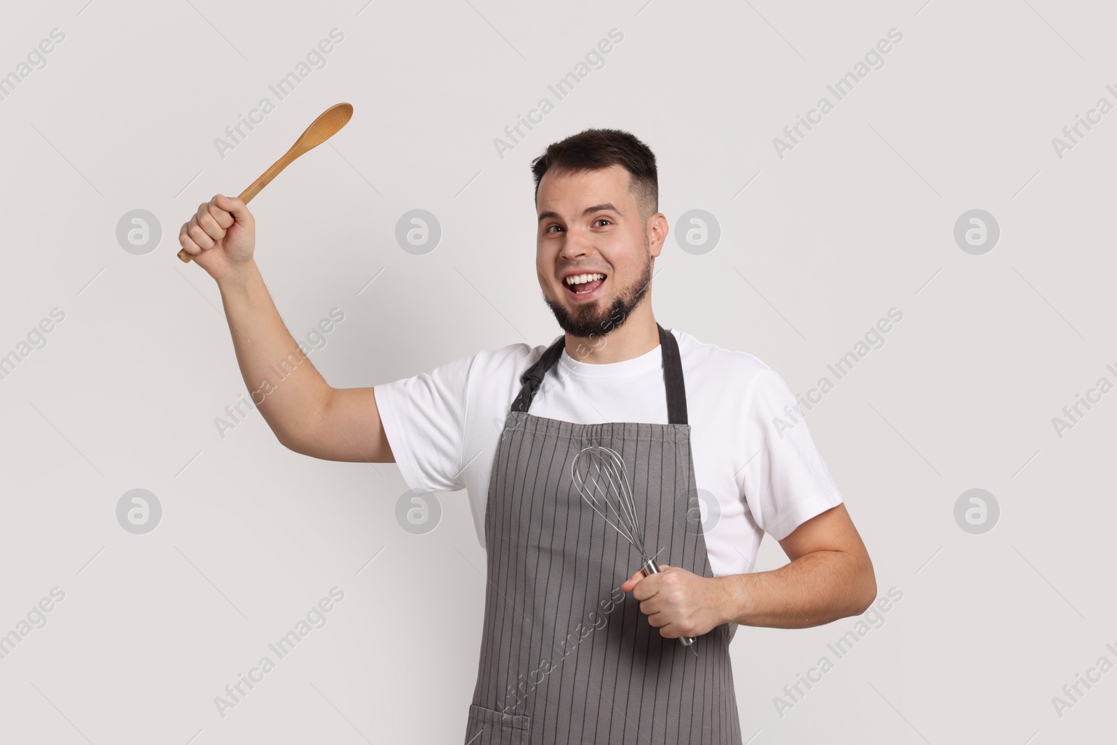 Photo of Happy professional confectioner in apron holding whisk and spoon on light grey background