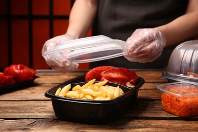 Waiter in gloves closing container with fresh prepared meal at wooden table, closeup. Food delivery service