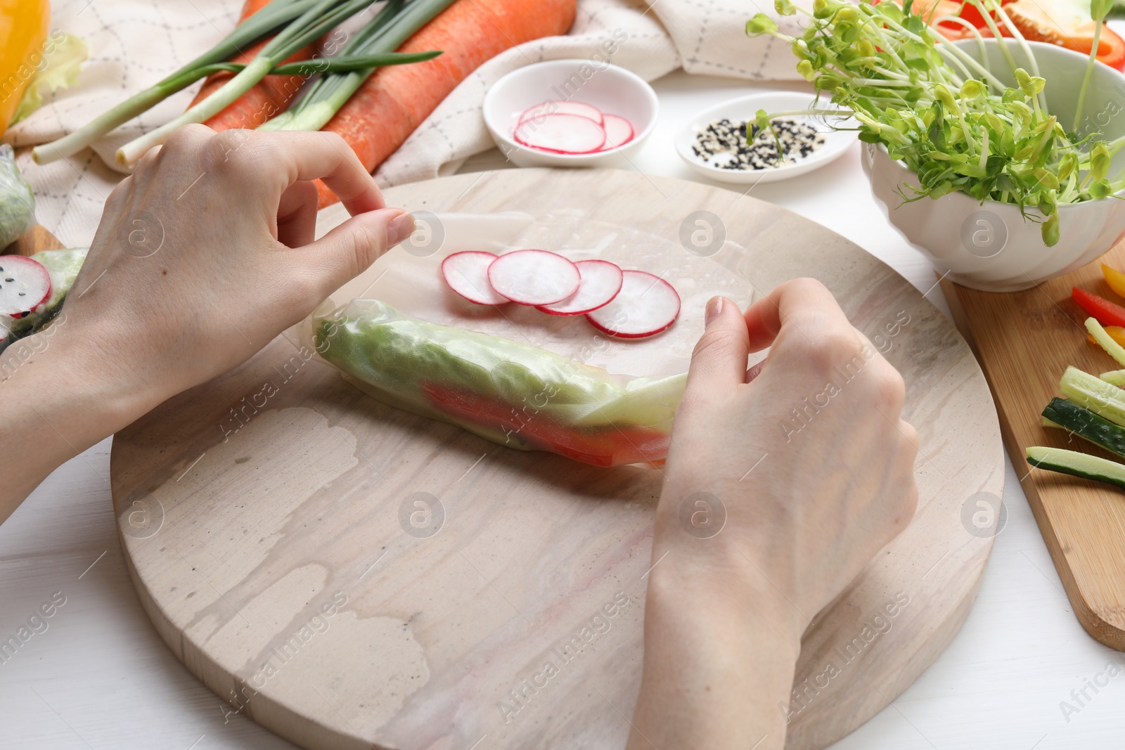 Photo of Woman wrapping spring roll at white wooden table with products, closeup