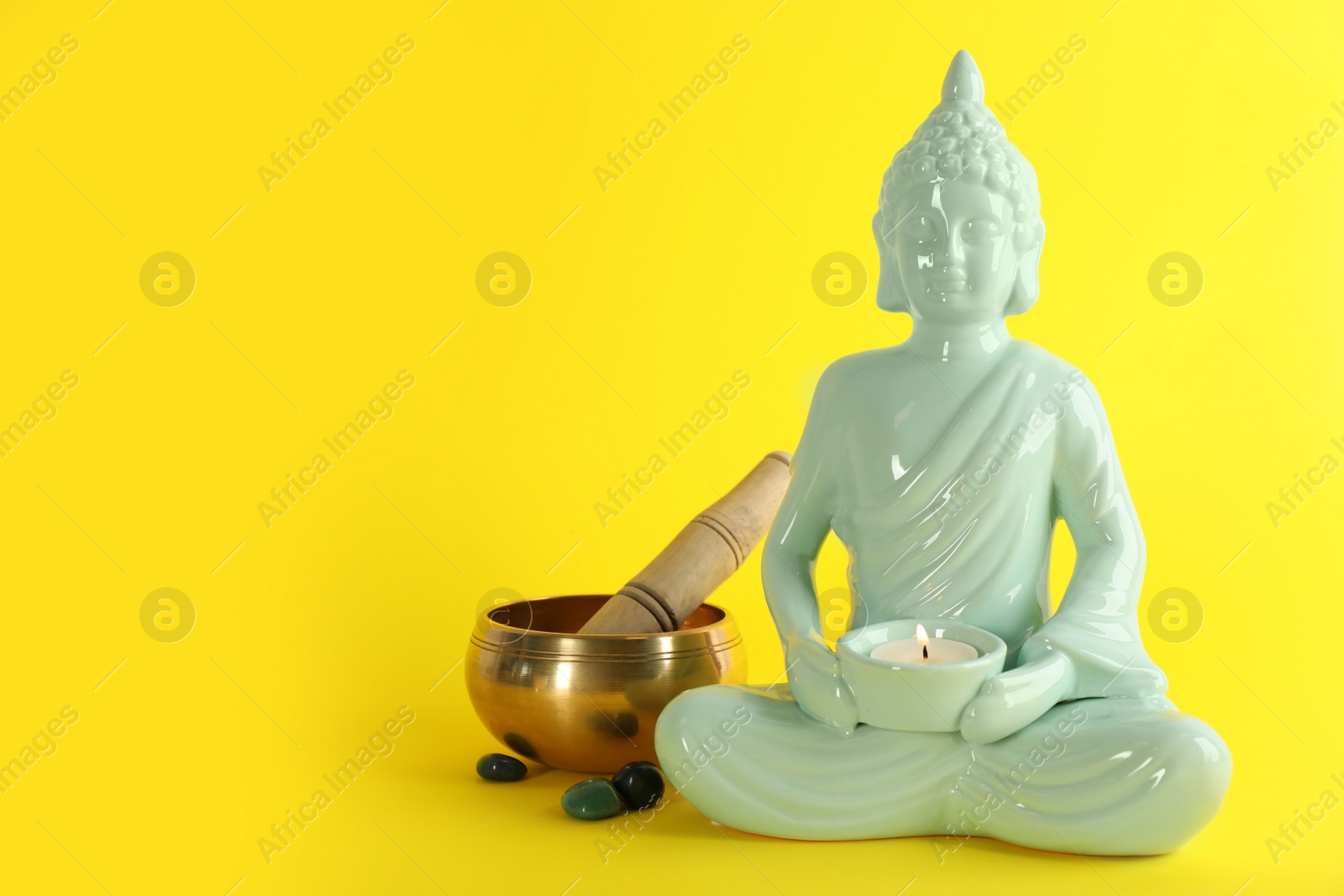Photo of Beautiful ceramic Buddha sculpture with burning candle and singing bowl on yellow background. Space for text