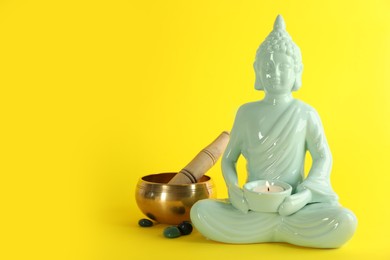 Beautiful ceramic Buddha sculpture with burning candle and singing bowl on yellow background. Space for text