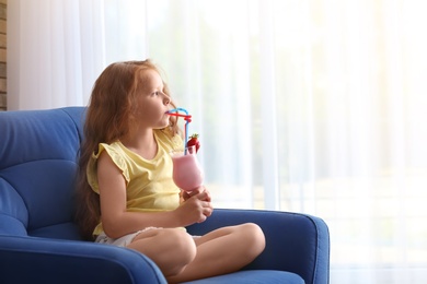 Little girl with glass of delicious milk shake in armchair indoors