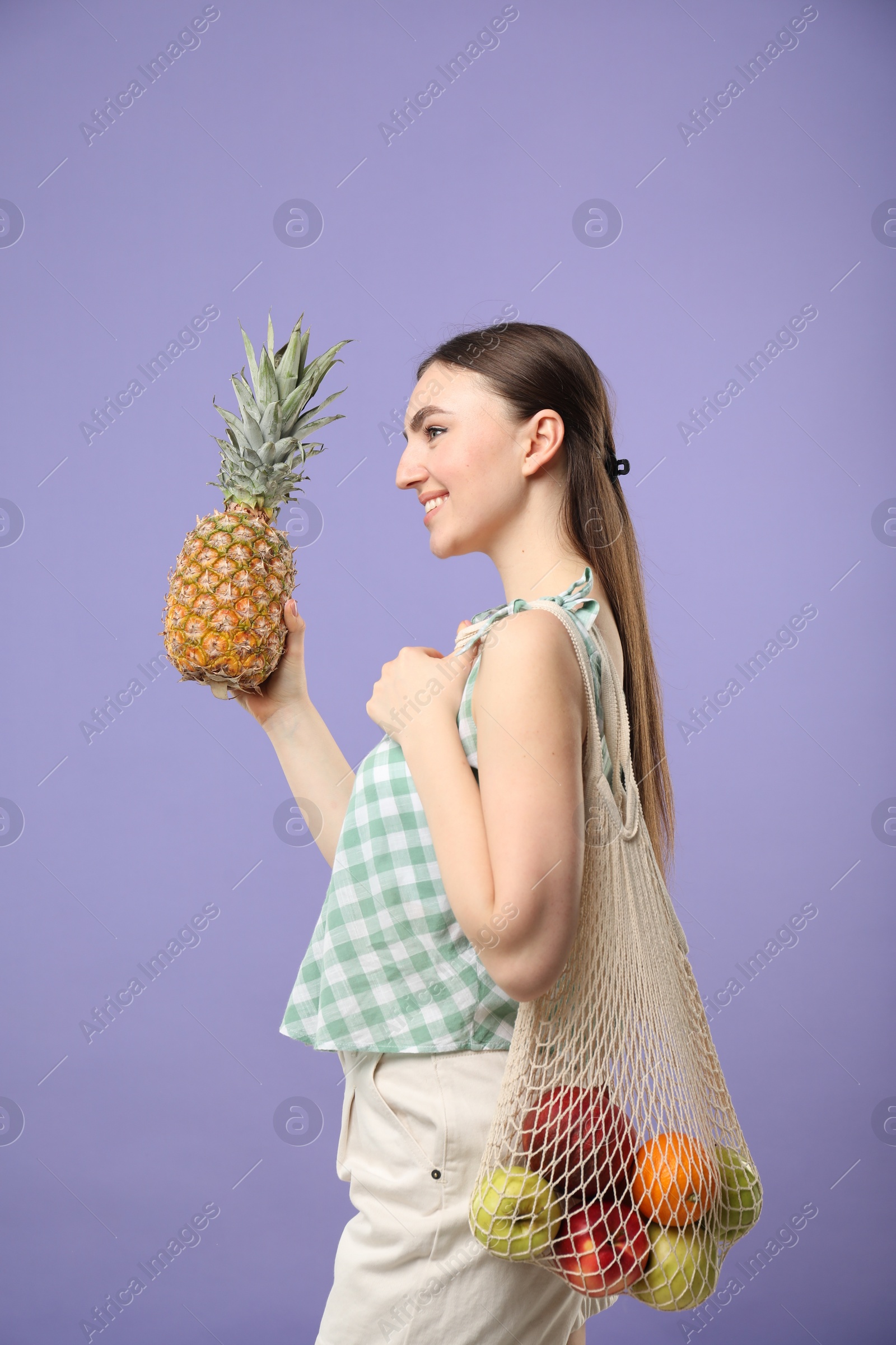 Photo of Woman with string bag of fresh fruits holding pineapple on violet background