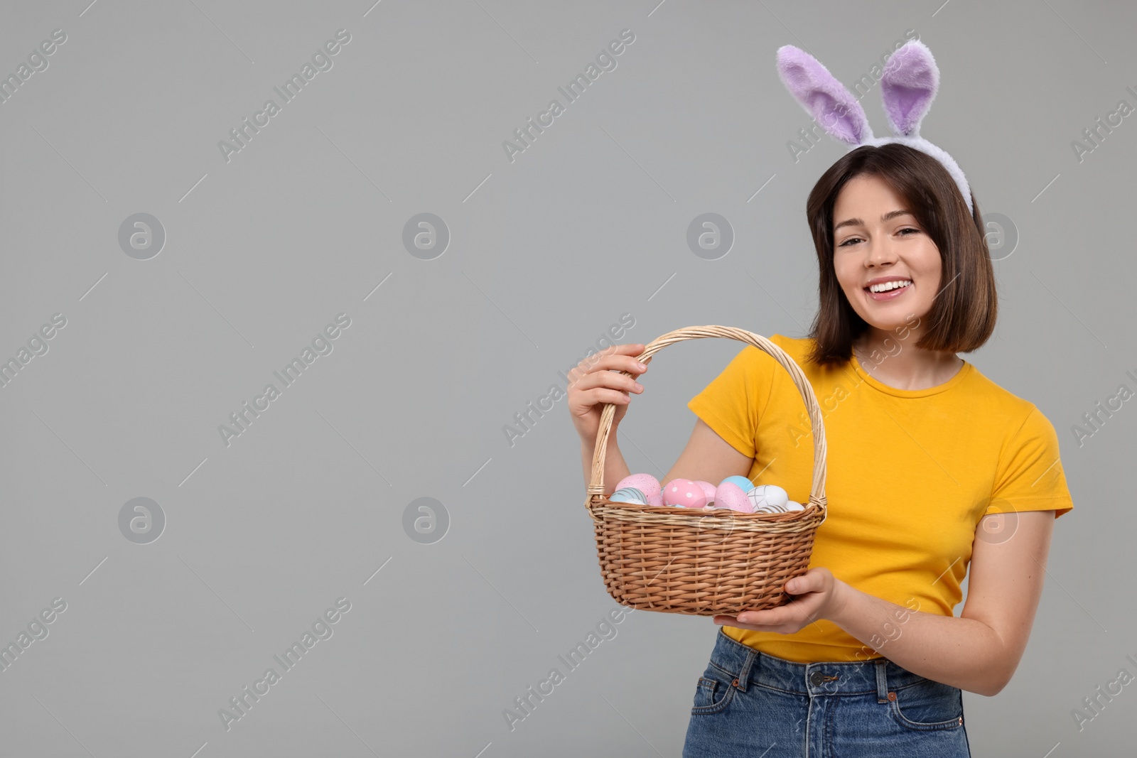 Photo of Easter celebration. Happy woman with bunny ears and wicker basket full of painted eggs on grey background, space for text