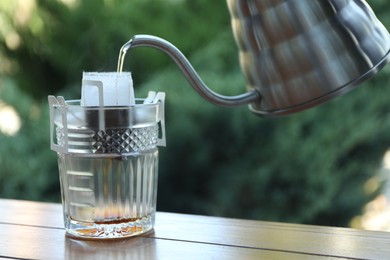 Pouring hot water into glass with drip coffee bag from kettle on wooden table, closeup