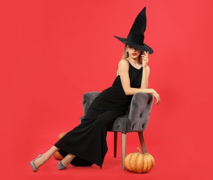 Photo of Beautiful woman wearing witch costume in armchair and pumpkins on red background, space for text. Halloween party