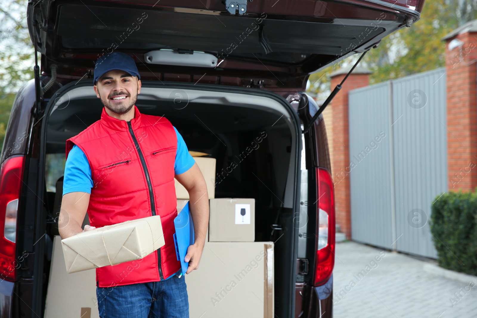 Photo of Young courier with parcel and clipboard near delivery van outdoors