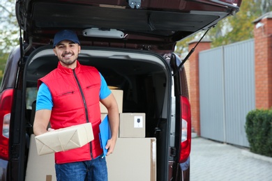 Young courier with parcel and clipboard near delivery van outdoors