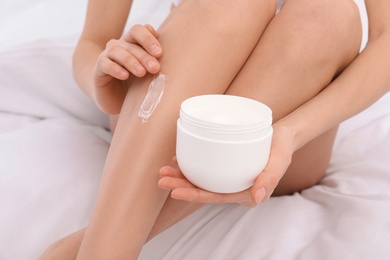 Photo of Young woman applying cream on her legs on bed, closeup. Beauty and body care