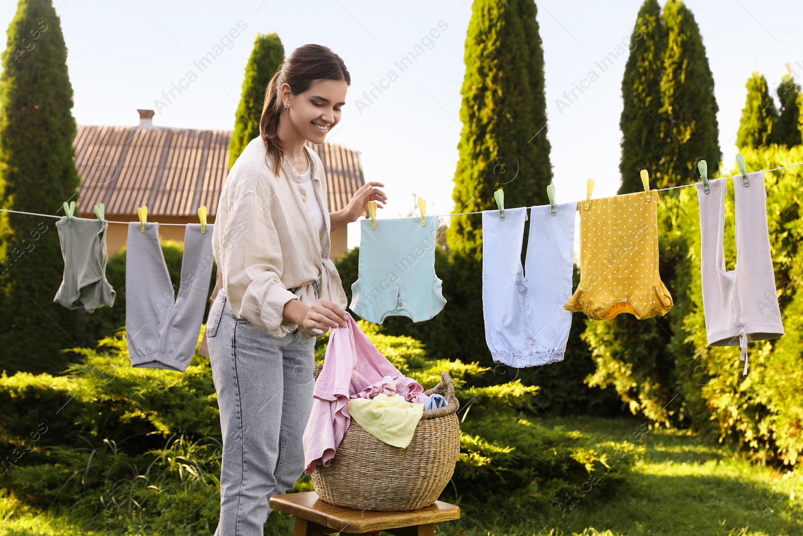 Photo of Smiling woman hanging baby clothes with clothespins on washing line for drying in backyard