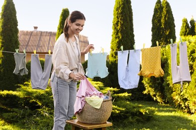 Photo of Smiling woman hanging baby clothes with clothespins on washing line for drying in backyard