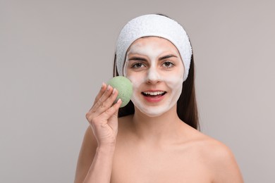 Young woman with headband washing her face using sponge on light grey background