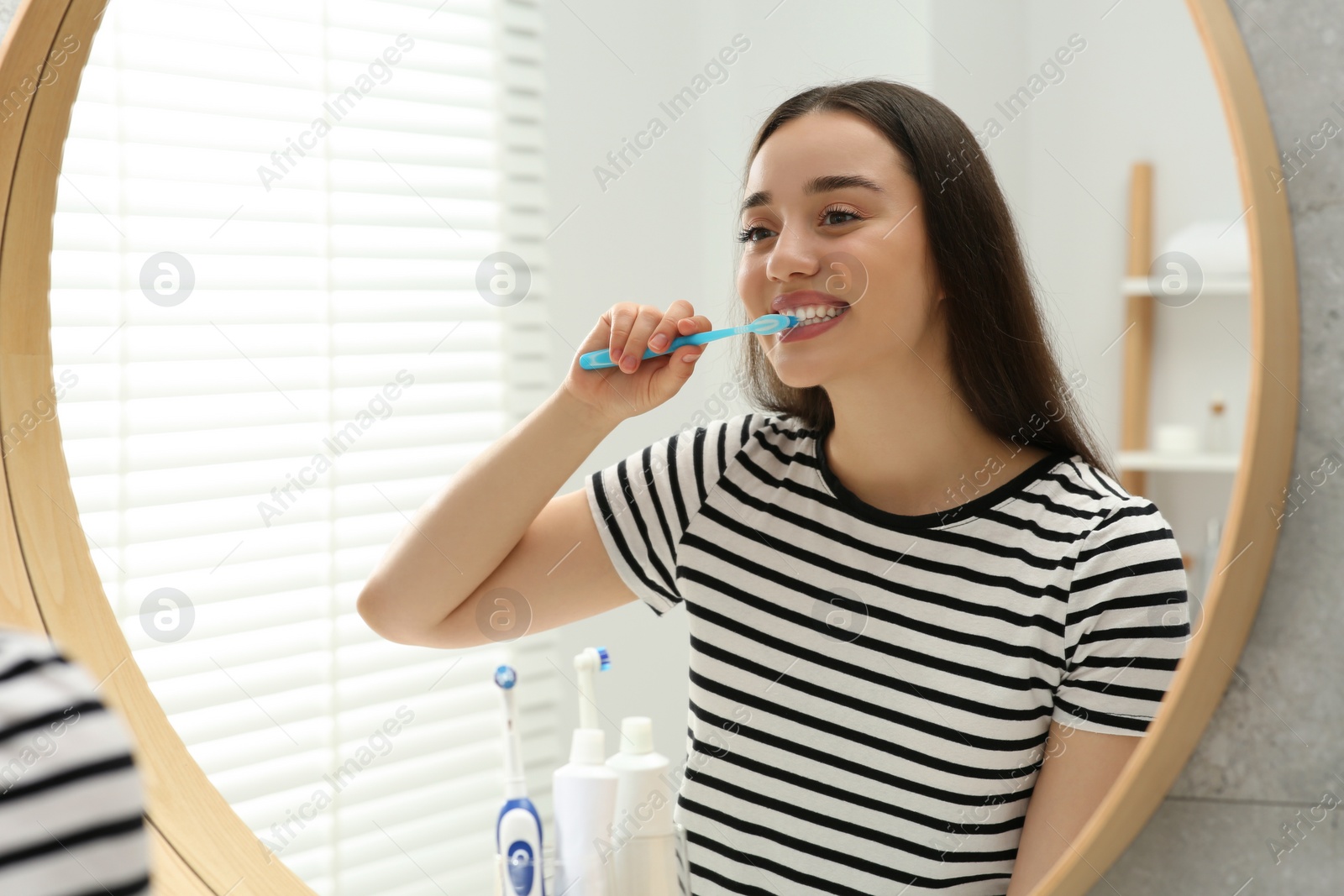 Photo of Young woman brushing her teeth with plastic toothbrush near mirror in bathroom
