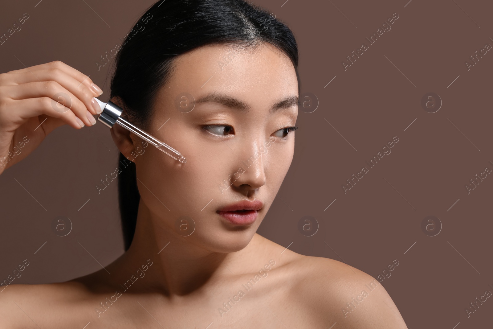 Photo of Beautiful young woman applying cosmetic serum onto her face on brown background, space for text