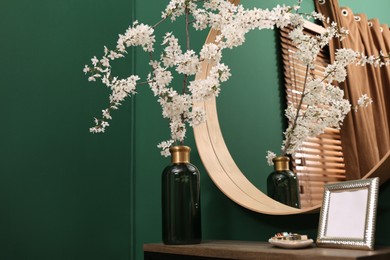 Photo of Blossoming tree twigs in vase and empty photo frame on wooden table near mirror indoors