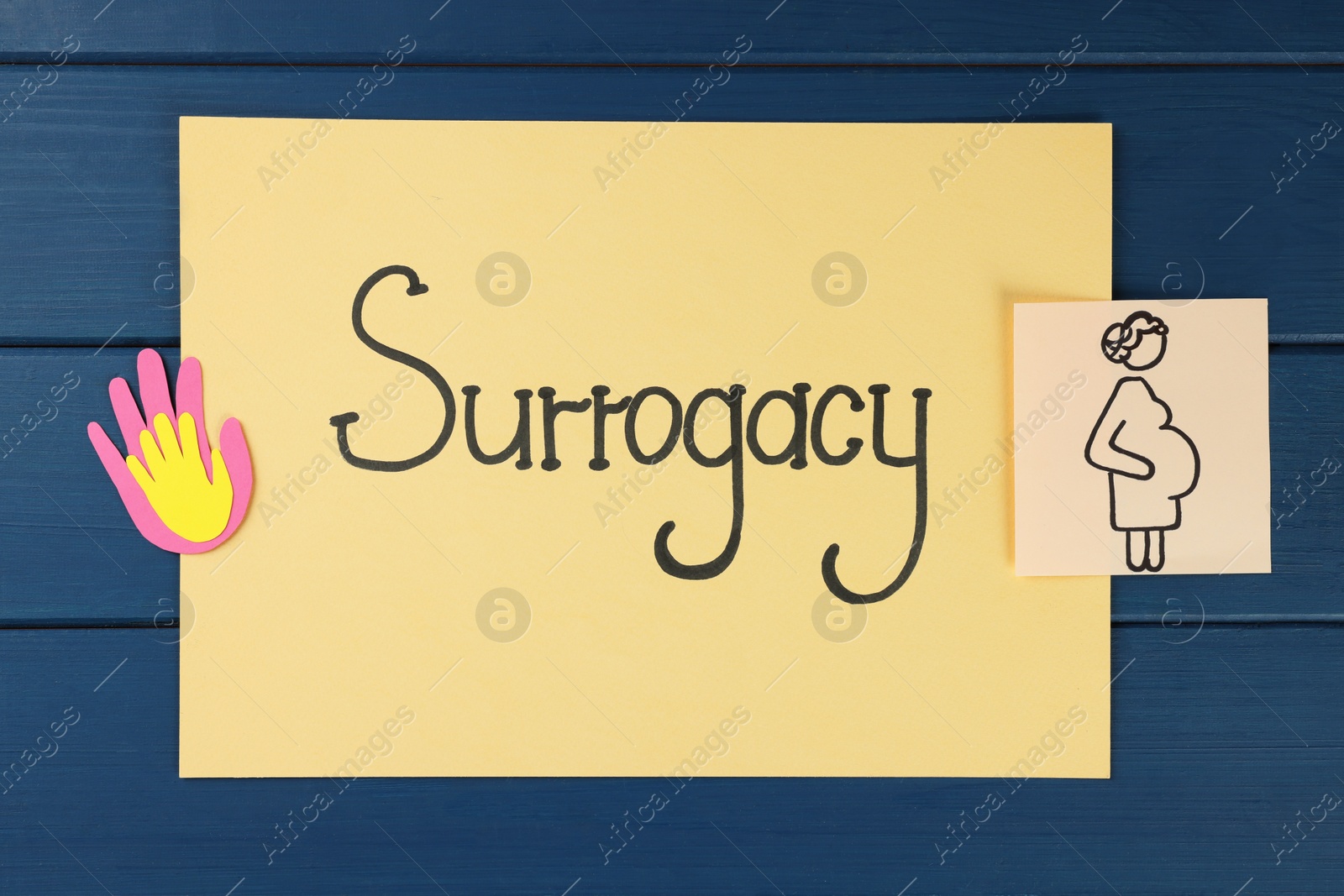 Photo of Sheet with word Surrogacy, paper palms and drawing of pregnant woman on blue wooden table, flat lay