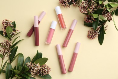 Different lip glosses and flowers on pale yellow background, flat lay