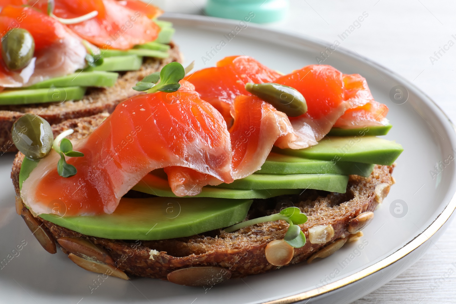 Photo of Delicious sandwich with salmon, avocado and capers on plate, closeup