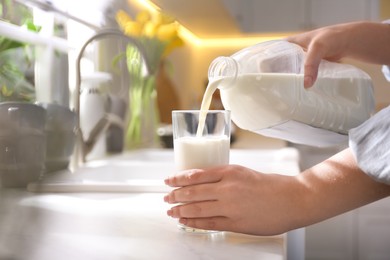 Photo of Young woman pouring milk from gallon bottle into glass at white countertop in kitchen, closeup