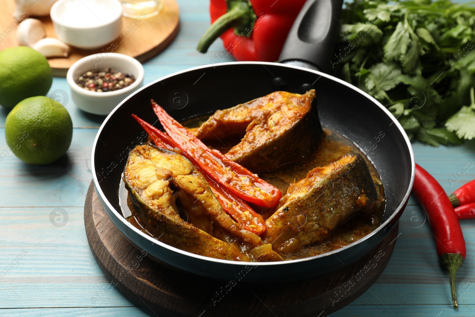 Photo of Tasty fish curry in frying pan and ingredients on light blue wooden table, closeup. Indian cuisine