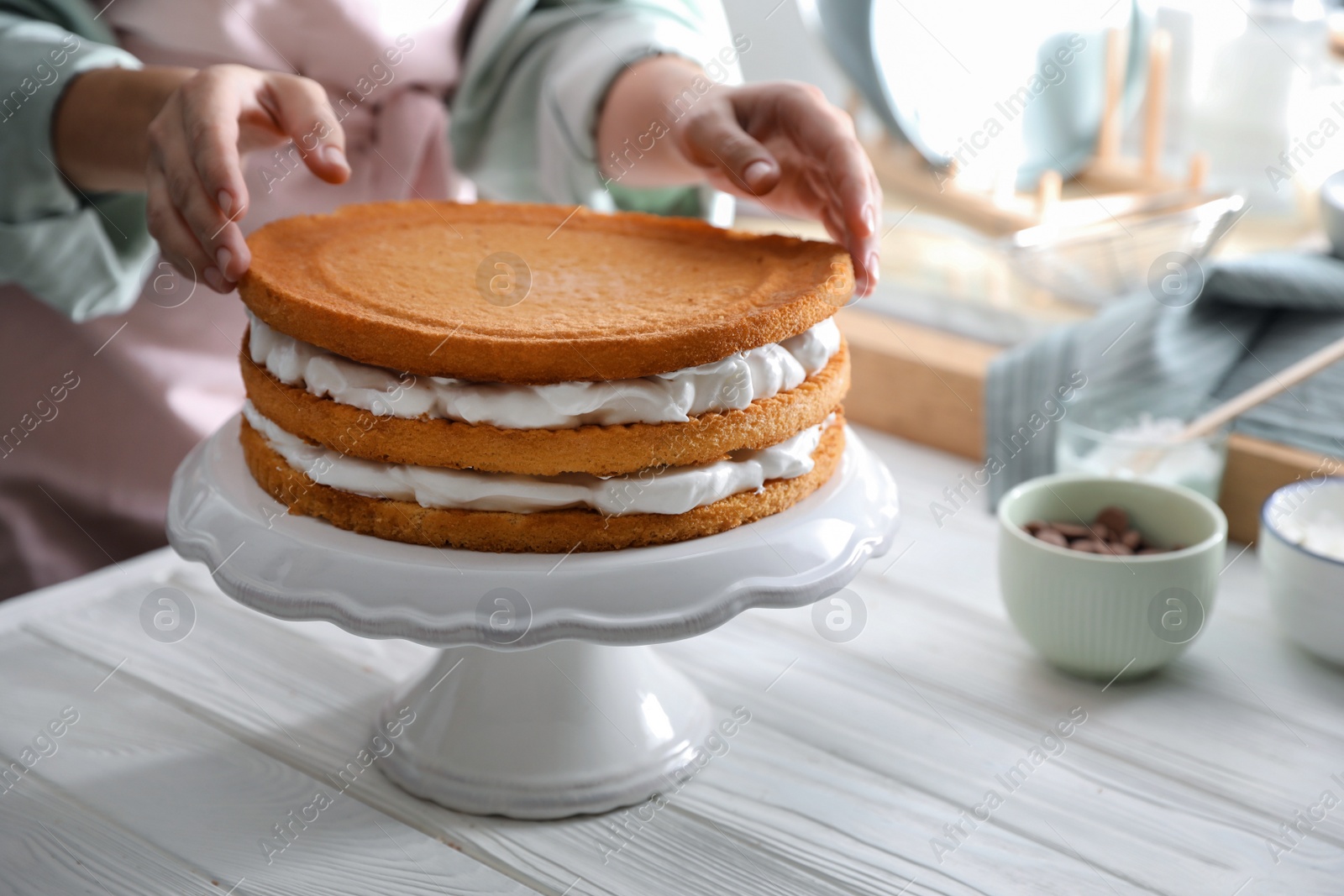 Photo of Woman stacking homemade sponge cakes at white wooden table, closeup