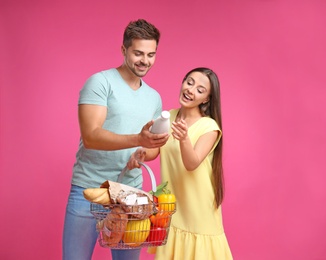 Young couple with shopping basket full of products on pink background