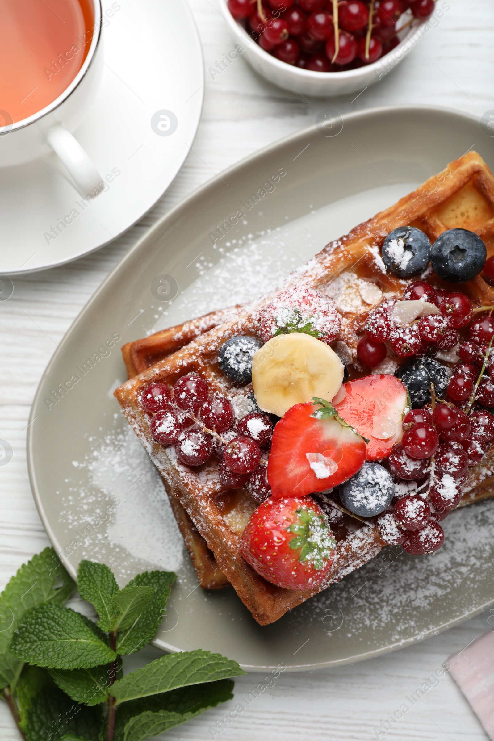 Photo of Plate of delicious Belgian waffles with berries, banana and powdered sugar on white wooden table, flat lay