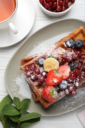 Photo of Plate of delicious Belgian waffles with berries, banana and powdered sugar on white wooden table, flat lay